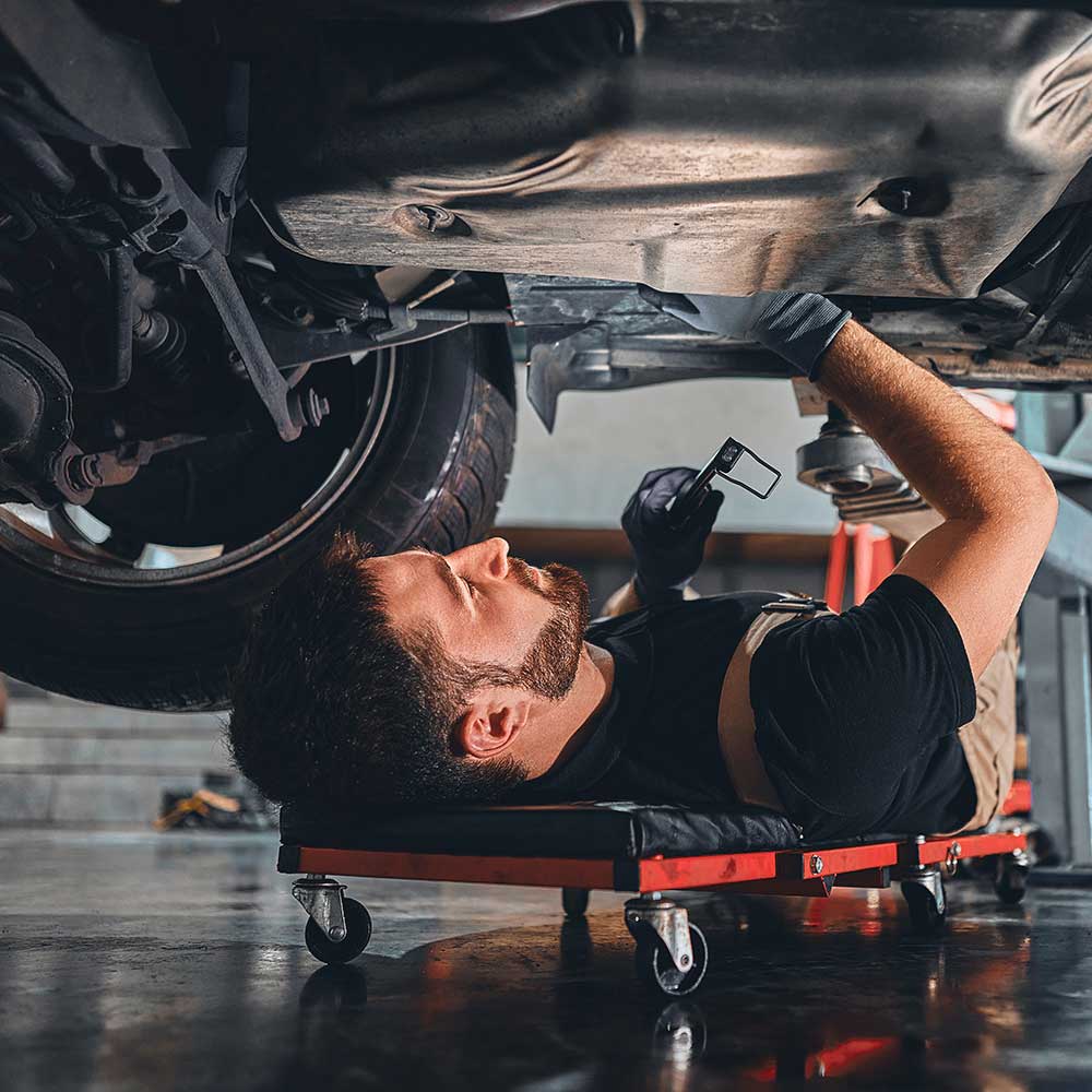 Männlicher Automechaniker, der mit einem Schraubenschlüssel für Reparaturen und Wartungsarbeiten unter dem Auto arbeitet. Mechaniker-Fahrzeugservice überprüft unter dem Auto in der Garage.